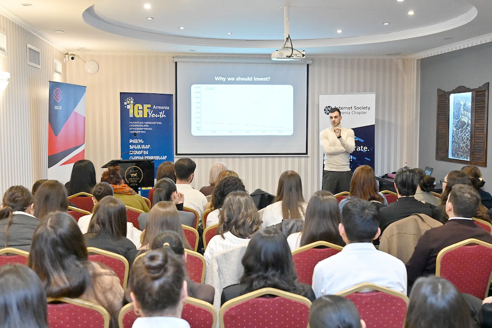 A man in a white sweater stands and speaks to a seated crowd