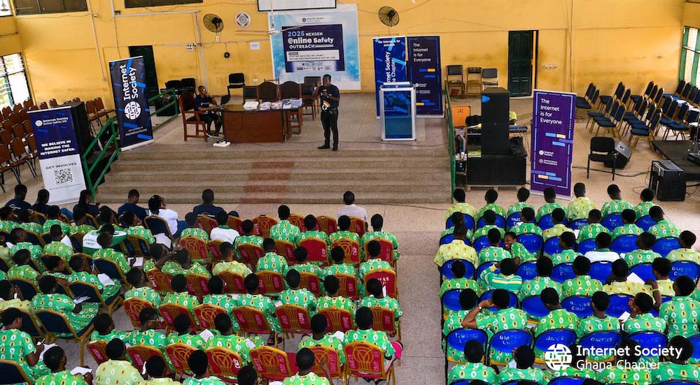 A man stands and speaks to a seated group of people wearing green shirts. Behind him, a sign says: 2025 NextGen Online Safety Outreach