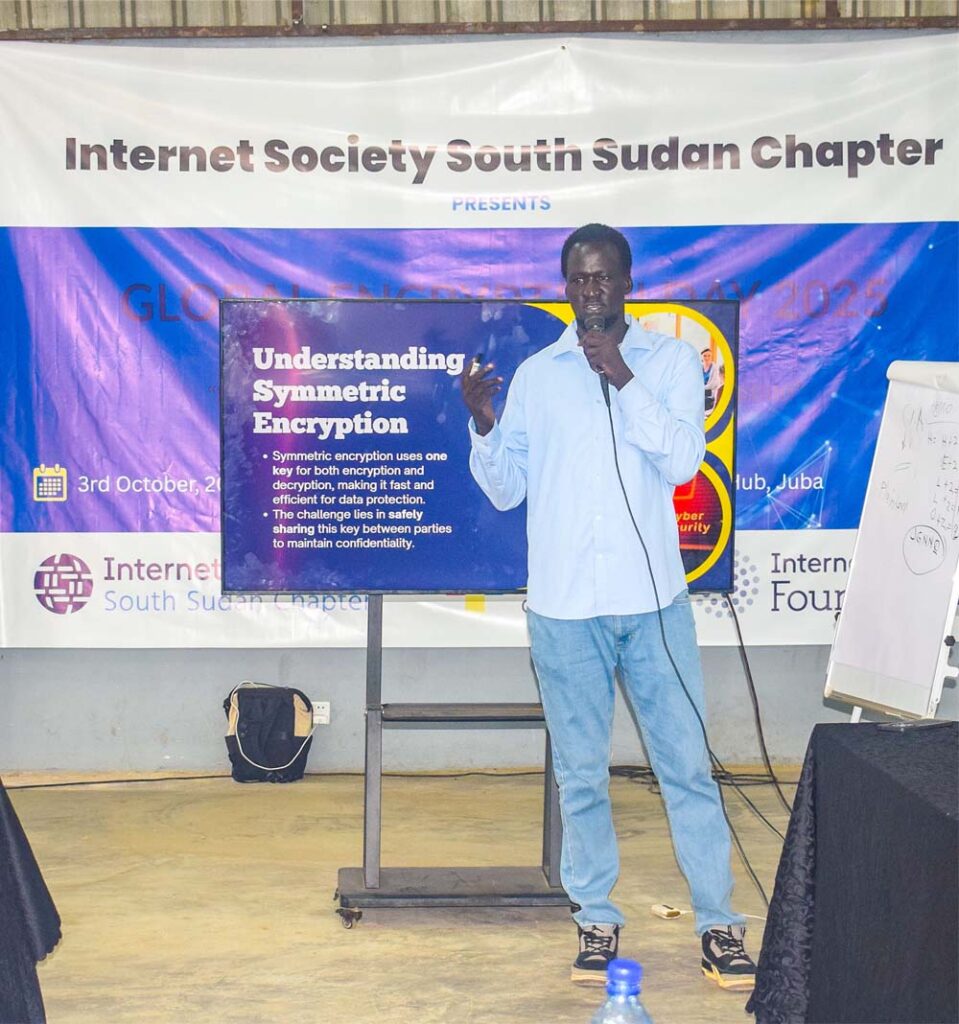 A man stands and talks into a microphone. A sign behind him says Internet Society South Sudan Chapter