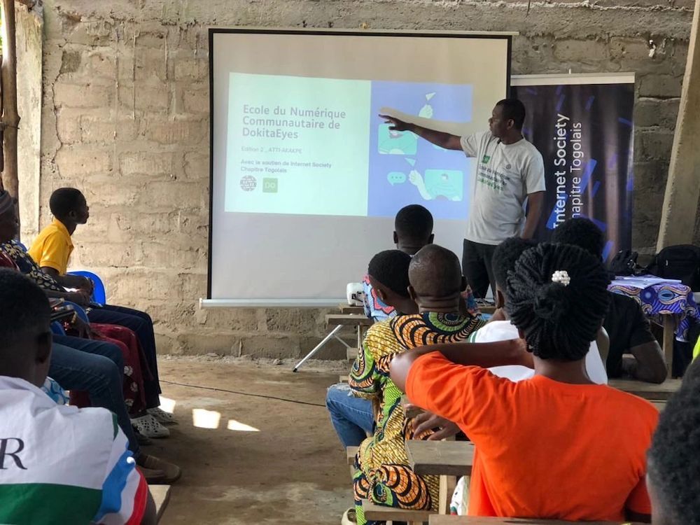 A man stands next to a projector screen and speaks to a seated group at a Togo Chapter digital literacy event